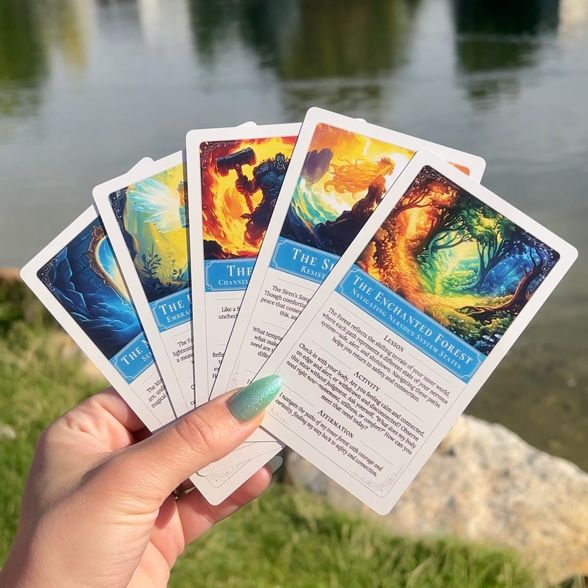 Hand holding self-discovery cards with fantasy-themed artwork against a sunny natural pond in the background