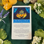 A card titled 'The Rebel's Mask' is displayed flat on a dark green background decorated with leaves, white flowers, crystals, and a gold ornament. The card features an illustration of a face wearing a blue and gold mask, surrounded by text instructions.