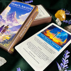 A boxed oracle card deck titled "Hero's Ascent" shown with one card laid out on a dark green surface. The box and card feature fantasy artwork with blue, gold, orange, and purple tones, and the scene is styled with crystals and purple flowers.