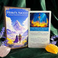 The Hero's Ascent card deck set displayed with the illustrated box, a sample card titled 'The Hero's Sword,' and decorative crystals on a dark green fabric background.