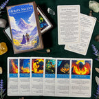 A Hero's Ascent card deck is displayed on a dark green cloth with several illustrated cards spread out in front. The box shows fantasy artwork of a mountain landscape with two figures, and the cards feature colorful scenes such as fire, storm, forest, and dragon imagery.